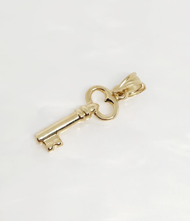 May include: A gold-colored key pendant with a classic design. The key features a detailed bow and a textured shaft, suggesting a vintage or antique aesthetic. The pendant is likely made of a precious metal, such as gold, and is designed to be worn as jewelry.