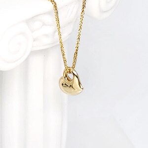 May include: A gold-coloured necklace featuring a small, smooth, heart-shaped pendant suspended from a delicate, woven chain. The pendant has a polished, reflective surface and a simple, rounded design that hangs vertically from the chain link.
