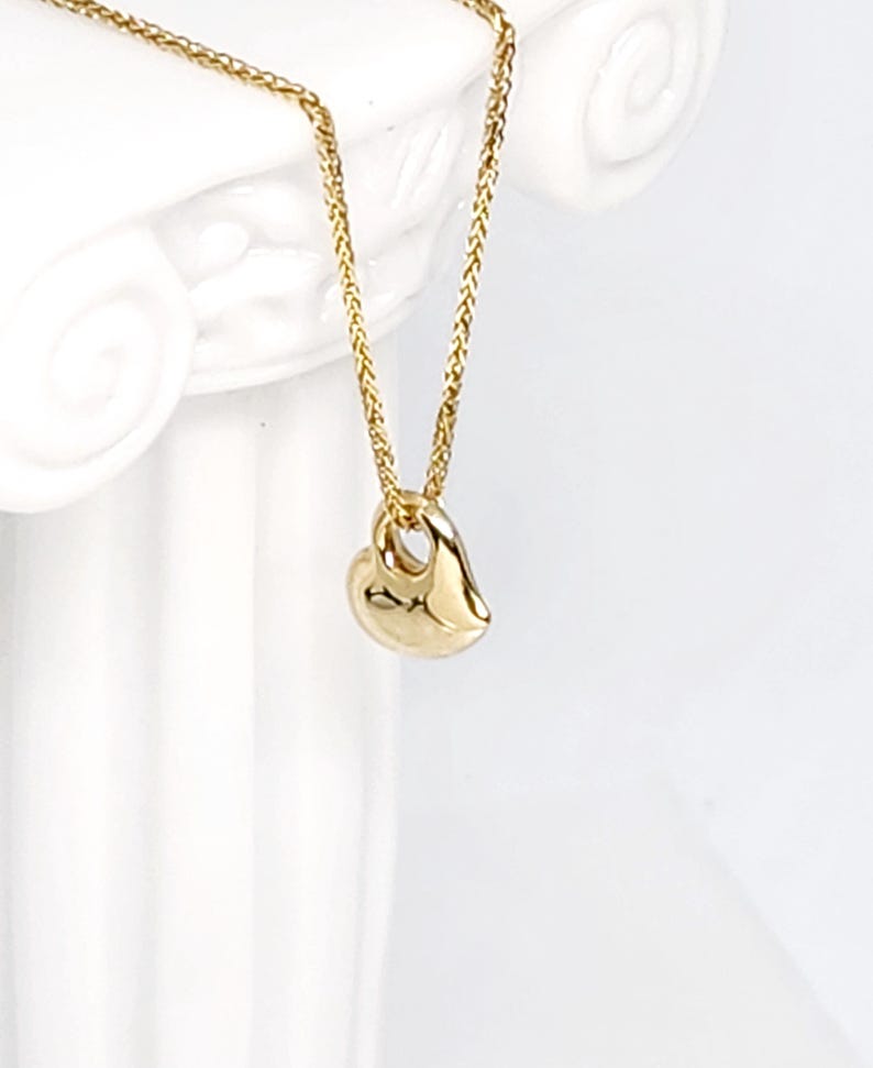 May include: A gold-coloured necklace featuring a small, smooth, heart-shaped pendant suspended from a delicate, woven chain. The pendant has a polished, reflective surface and a simple, rounded design that hangs vertically from the chain link.