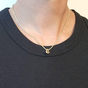 May include: A thin gold chain necklace featuring a small, circular gold pendant. The necklace rests against a black ribbed shirt. The chain has a delicate, fine link construction, and the pendant is simple and round in shape, reflecting light.