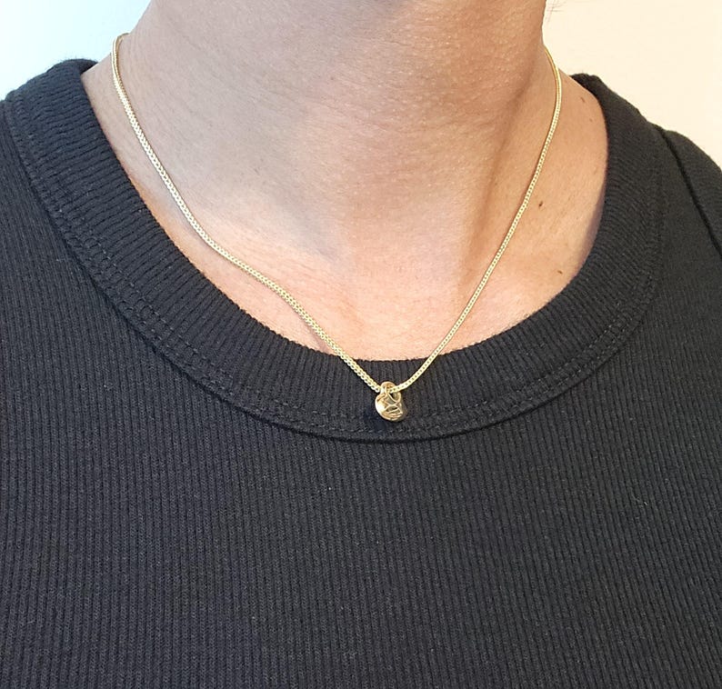 May include: A thin gold chain necklace featuring a small, circular gold pendant. The necklace rests against a black ribbed shirt. The chain has a delicate, fine link construction, and the pendant is simple and round in shape, reflecting light.