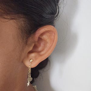 May include: Gold dangle earrings with a heart and star charm. The earrings are hanging from a chain.