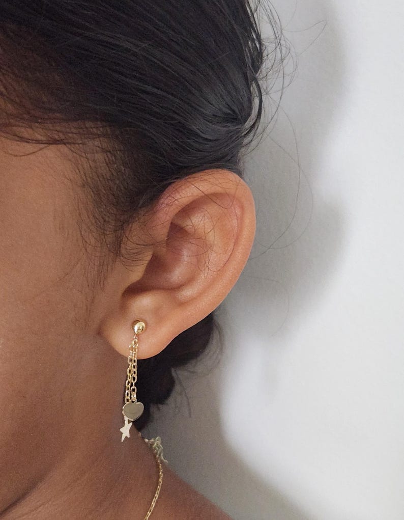 May include: Gold dangle earrings with a heart and star charm. The earrings are hanging from a chain.