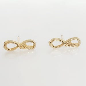 May include: A pair of gold-colored stud earrings. Each earring is in the shape of an infinity symbol with the word "love" inscribed on the lower loop. The earrings are set against a white background.