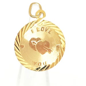 May include: A round gold pendant featuring a textured border and a central design of two hearts pierced by an arrow. The text I LOVE YOU is engraved on the face of the pendant. It includes a small circular gold bail at the top for attachment.