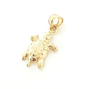 May include: A gold-toned turtle pendant featuring a detailed shell pattern and four flippers. The pendant includes a small, rounded bail at the top for attachment to a chain, showcasing a polished, reflective surface throughout the entire piece.