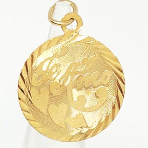 May include: A round gold pendant featuring a textured surface with a diagonal striped border. The center displays the cursive script text 'Selma' alongside two heart shapes. The pendant includes a small circular jump ring at the top for attachment.