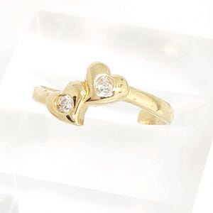 May include: A gold ring featuring two adjacent heart-shaped settings, each containing a single clear round gemstone. The band is polished gold and has an open design, showcasing the two hearts positioned side by side at the center of the ring.