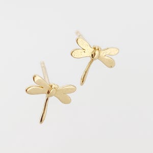 May include: A pair of gold-colored dragonfly stud earrings. Each earring features a detailed dragonfly design with delicate wings and a slender body. The earrings are set against a plain white background, highlighting their intricate details.