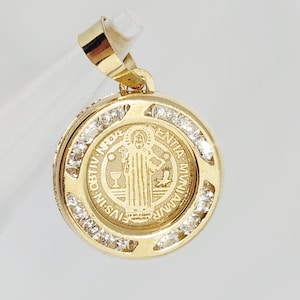 Puede incluir: Colgante dorado con diseño redondo. El colgante presenta un grabado detallado de una figura religiosa y está rodeado por una fila de pequeñas piedras preciosas transparentes. La argolla también es dorada. El colgante es probablemente de oro.