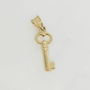 May include: A gold-colored key pendant with a detailed design. The key features a decorative bow, a long stem, and a detailed bit. The pendant is likely made of gold and is designed to be worn as jewelry.