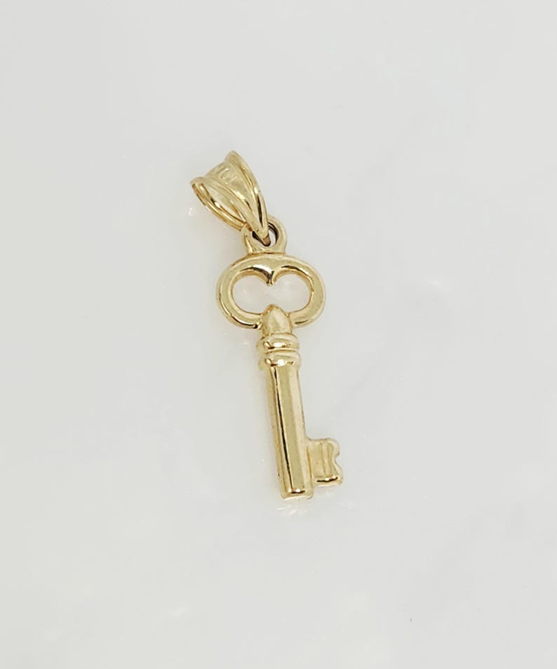 May include: A gold-colored key pendant with a detailed design. The key features a decorative bow, a long stem, and a detailed bit. The pendant is likely made of gold and is designed to be worn as jewelry.
