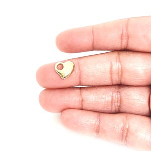 May include: A small, polished gold-tone heart-shaped charm featuring a single circular hole at the top for attachment. The charm has a smooth, reflective surface and a rounded, symmetrical design, presented as a singular jewellery component.