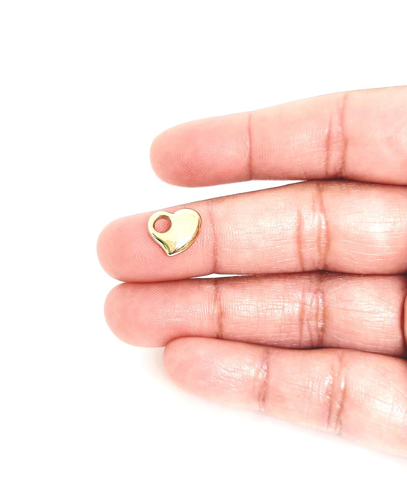 May include: A small, polished gold-tone heart-shaped charm featuring a single circular hole at the top for attachment. The charm has a smooth, reflective surface and a rounded, symmetrical design, presented as a singular jewellery component.