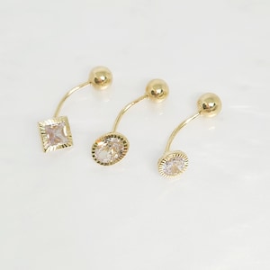 Puede incluir: Tres anillos de ombligo de oro con piedras de cristal transparente. Las piedras son un cuadrado, un círculo y un círculo con un diámetro ligeramente más pequeño.