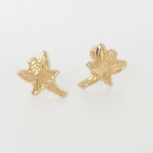 Puede incluir: Un par de pendientes dorados. Cada pendiente tiene la forma de un peque&ntilde;o hada con alas extendidas y una cara detallada. Los pendientes est&aacute;n sobre un fondo blanco liso.