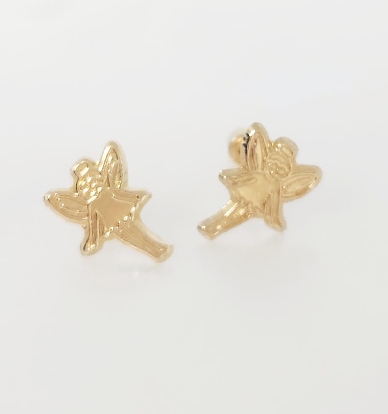 Puede incluir: Un par de pendientes dorados. Cada pendiente tiene la forma de un peque&ntilde;o hada con alas extendidas y una cara detallada. Los pendientes est&aacute;n sobre un fondo blanco liso.