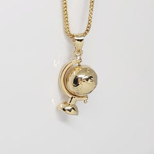 May include: A gold-coloured pendant necklace featuring a globe charm. The globe is mounted on a stand and suspended from a chain. The pendant and chain are made of a shiny, gold-toned metal.