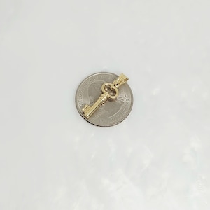 May include: A gold-colored key pendant with a detailed design, resting on a quarter. The key has a classic skeleton key shape with a loop at the top for a chain. The pendant is likely made of gold.