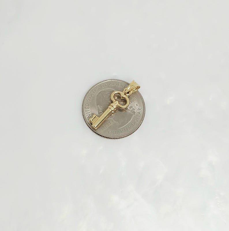May include: A gold-colored key pendant with a detailed design, resting on a quarter. The key has a classic skeleton key shape with a loop at the top for a chain. The pendant is likely made of gold.