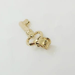 May include: A gold-colored key pendant with a decorative rose design at the end. The key has a classic shape with a loop and a detailed bow. The pendant is likely made of metal and has a shiny finish.