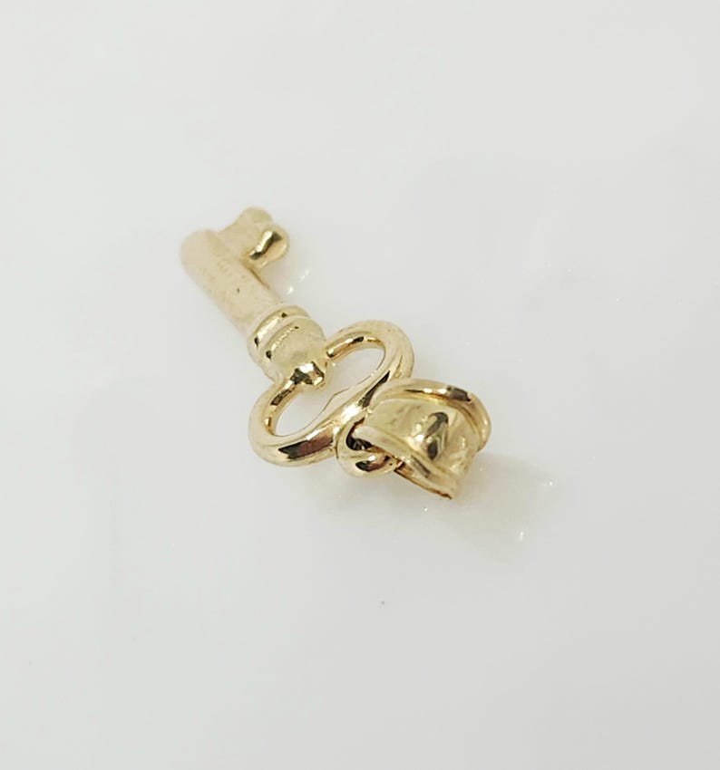 May include: A gold-colored key pendant with a decorative rose design at the end. The key has a classic shape with a loop and a detailed bow. The pendant is likely made of metal and has a shiny finish.