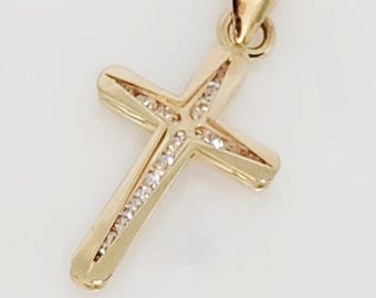 May include: A gold-colored cross pendant with a loop for a chain. The cross features a series of small, clear gemstones set along the arms and at the center. The pendant is likely made of gold and is displayed against a white background.