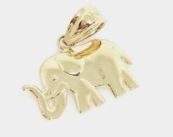 May include: A polished gold-tone metal pendant shaped like an elephant in profile, featuring a smooth, reflective surface and a large, integrated bail at the top for attachment to a chain or cord, showcasing a classic, simple silhouette design.