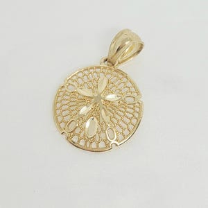 May include: A gold-coloured sand dollar pendant. The pendant features an intricate openwork design with a central floral motif and oval cutouts. The bail has a textured finish.