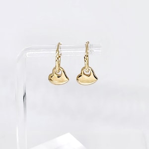 May include: A pair of gold hoop earrings featuring small, smooth, polished heart-shaped charms. Each hoop has a textured, diamond-cut pattern along the upper portion, and the heart charms hang freely from the bottom of the circular gold hoops.