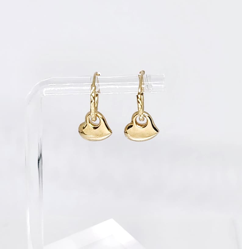 May include: A pair of gold hoop earrings featuring small, smooth, polished heart-shaped charms. Each hoop has a textured, diamond-cut pattern along the upper portion, and the heart charms hang freely from the bottom of the circular gold hoops.