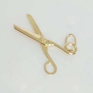 May include: A gold-colored pair of scissors, with open blades and finger holes. The scissors are set against a white background, and the blades are angled to the left. The scissors appear to be made of metal.