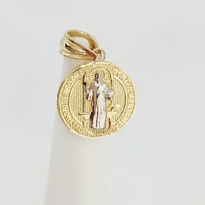 Puede incluir: Un colgante redondo de oro con una figura central plateada de un santo sosteniendo un bastón. El fondo del colgante tiene un patrón de rejilla texturizado y el borde exterior contiene texto en latín en relieve. Incluye una anilla de oro.