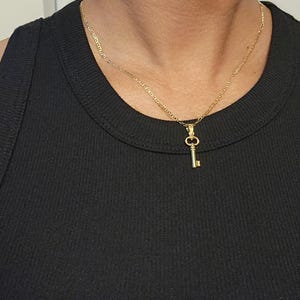 May include: A gold-colored key pendant necklace. The key charm is small and detailed, hanging from a delicate chain. The necklace is worn against a black top, creating a contrast that highlights the jewelry.