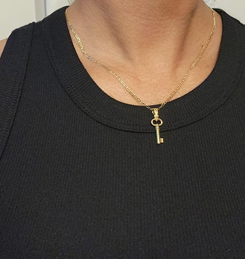 May include: A gold-colored key pendant necklace. The key charm is small and detailed, hanging from a delicate chain. The necklace is worn against a black top, creating a contrast that highlights the jewelry.