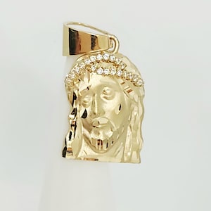 Op de afbeelding: Een gouden hanger met het gebeeldhouwde gezicht van Jezus en een doornenkroon bezet met kleine, heldere, fonkelende steentjes. De hanger heeft een gepolijst, reflecterend oppervlak en een grote, gladde, taps toelopende hangerlus aan de bovenkant.