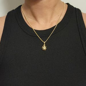 K&ouml;nnte beinhalten: Eine goldfarbene Halskette mit einer feinen Kette und einem kleinen, texturierten, abgerundeten Anh&auml;nger. Der Anh&auml;nger hat eine detaillierte, segmentierte Oberfl&auml;che, die an einen K&auml;fer oder ein Insekt erinnert, und h&auml;ngt mittig an der Kette.
