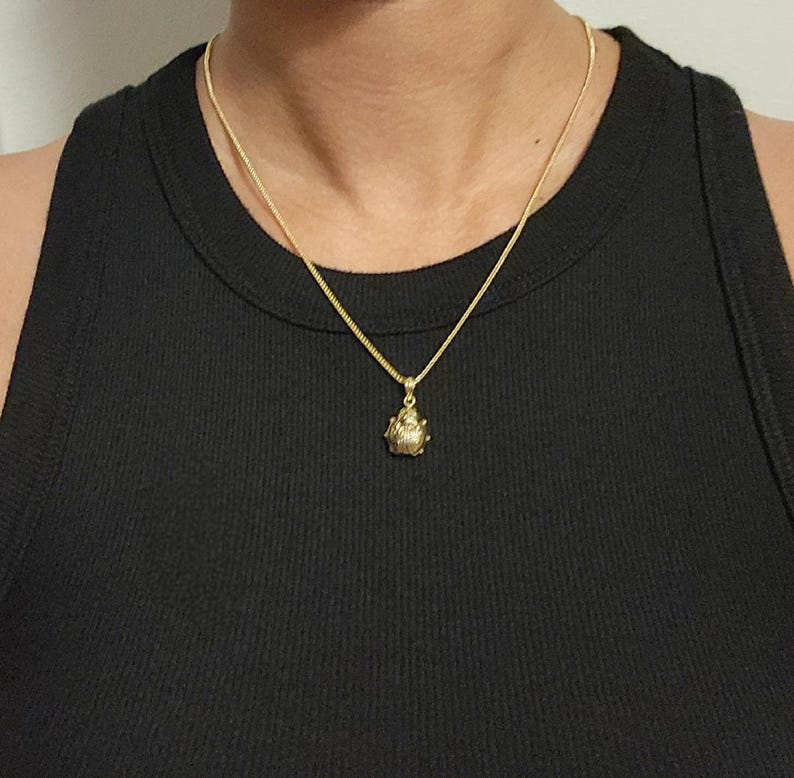 K&ouml;nnte beinhalten: Eine goldfarbene Halskette mit einer feinen Kette und einem kleinen, texturierten, abgerundeten Anh&auml;nger. Der Anh&auml;nger hat eine detaillierte, segmentierte Oberfl&auml;che, die an einen K&auml;fer oder ein Insekt erinnert, und h&auml;ngt mittig an der Kette.
