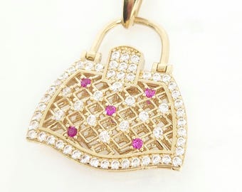 May include: A gold-toned pendant shaped like a handbag, featuring a lattice-patterned body encrusted with clear white stones and accented by five small, round, vibrant pink stones. The piece includes a bail at the top for attachment to a chain.