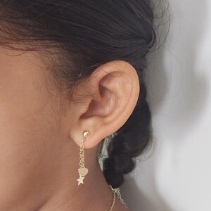 May include: Gold dangle earrings with a heart and star charm. The earrings are hanging from a person's ear.