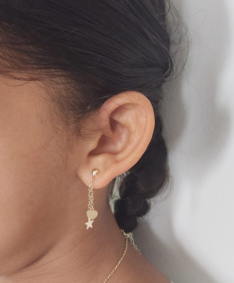 May include: Gold dangle earrings with a heart and star charm. The earrings are hanging from a person's ear.