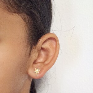 Puede incluir: Primer plano de una oreja con un peque&ntilde;o pendiente de bot&oacute;n dorado con forma de lib&eacute;lula. El pendiente presenta un dise&ntilde;o detallado de una lib&eacute;lula con las alas extendidas y un cuerpo delgado. El pendiente est&aacute; colocado contra la piel de la oreja.