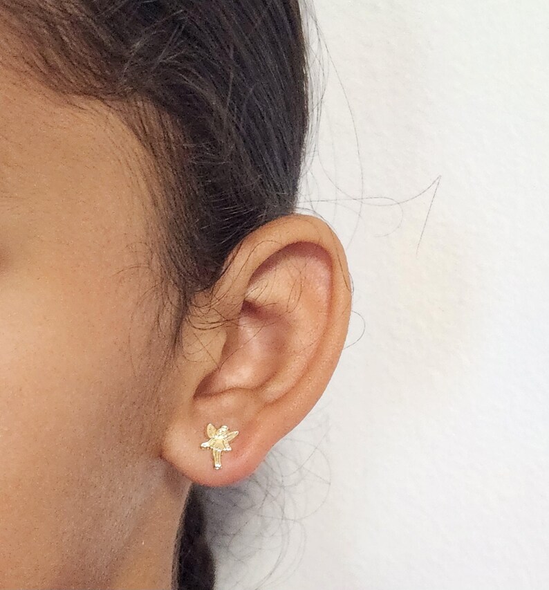 Puede incluir: Primer plano de una oreja con un peque&ntilde;o pendiente de bot&oacute;n dorado con forma de lib&eacute;lula. El pendiente presenta un dise&ntilde;o detallado de una lib&eacute;lula con las alas extendidas y un cuerpo delgado. El pendiente est&aacute; colocado contra la piel de la oreja.