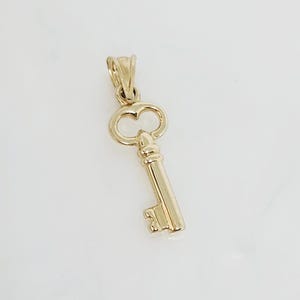 May include: A gold-colored key pendant with a classic design. The pendant features a detailed key shape, including the bow, the bit, and the stem. The key is made of a shiny, reflective metal, and is likely a charm or jewelry piece.