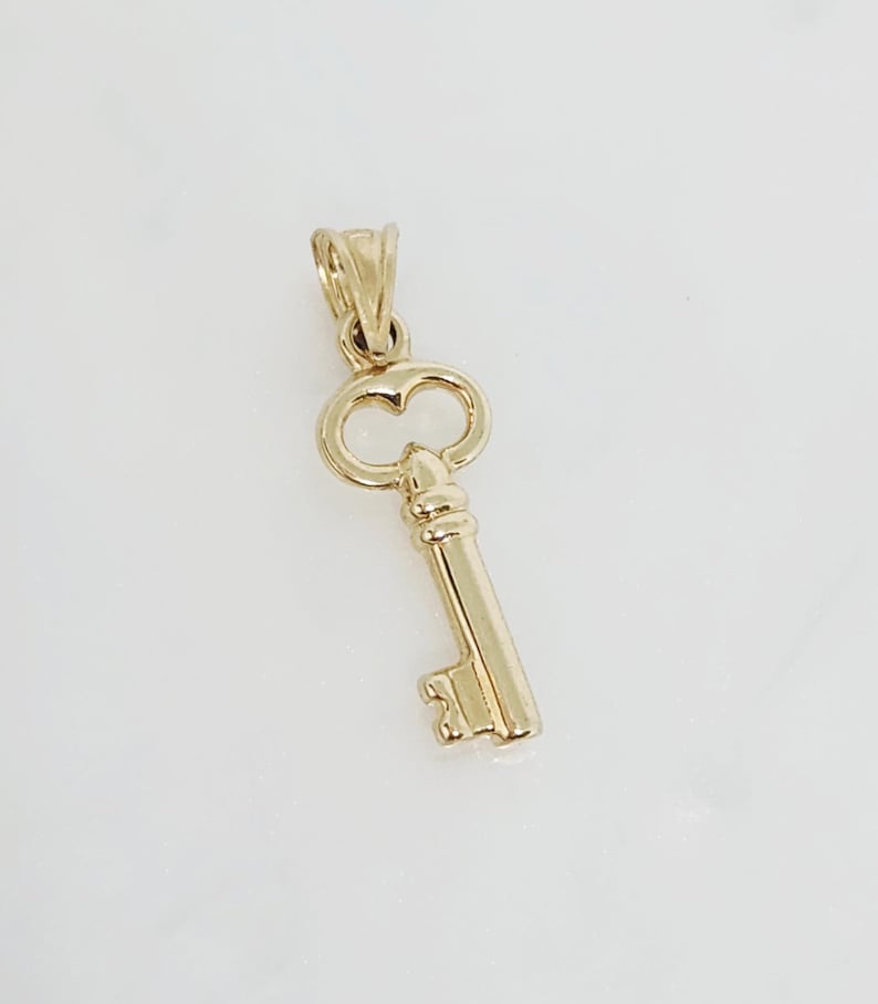 May include: A gold-colored key pendant with a classic design. The pendant features a detailed key shape, including the bow, the bit, and the stem. The key is made of a shiny, reflective metal, and is likely a charm or jewelry piece.