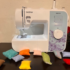 Puede incluir: Una máquina de coser Brother XM2701 blanca con un diseño floral en el lateral. La máquina está sobre una superficie marrón oscura con varios cuadrados pequeños de tela de colores.