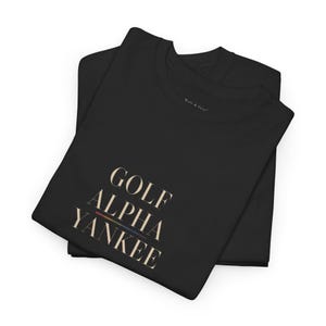 May include: Two black t-shirts are neatly folded and stacked. The top shirt features the words "GOLF ALPHA YANKEE" in a serif font, with a rainbow-colored stripe beneath the text. The brand name "Walt & Pete" is printed on the top right.