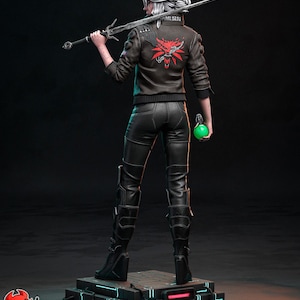 May include: A detailed figurine of a character from The Witcher series. The figure is wearing a black leather jacket with a red emblem on the back, black pants, and boots. She holds a sword and a green orb. The figure stands on a platform.