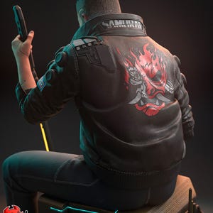 May include: A black leather jacket with the word "SAMURAI" on the collar and a red and gray graphic on the back. The jacket is worn by a person sitting on a box, holding a black device.