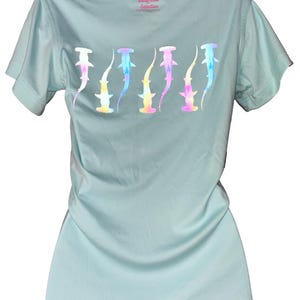 May include: A light blue t-shirt featuring a row of stylized hammerhead sharks in a rainbow holographic design. The shirt has short sleeves and a crew neck. The text "ArtOfficial Selection" is printed on the back of the neck.